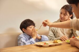 밥상 앞에서 손으로 입을 가리며 밥을 거부하는 아이와 먹이려는 엄마